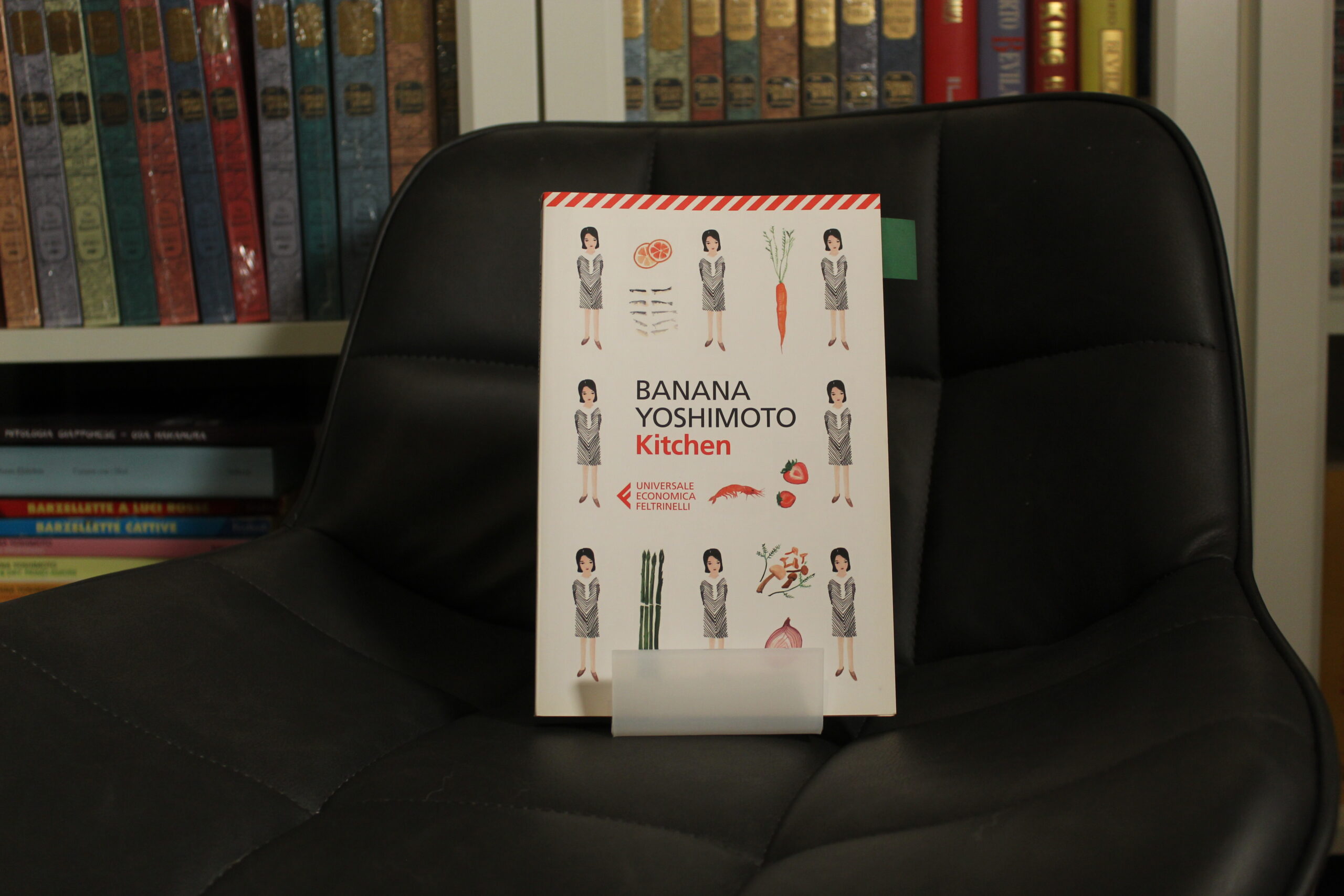 Copertina del libro Kitchen di Banana Yoshimoto edizione Feltrinelli appoggiata su una poltrona nera con una libreria sullo sfondo.